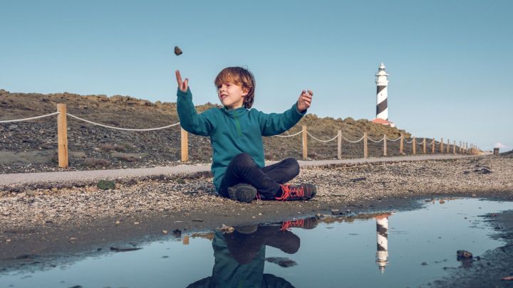 Ein Kind sitzt am Boden, wirft einen Stein in ein Pf&uuml;tze, w&auml;hrend ein Leuchtturm im Hintergrund sichtbar ist.