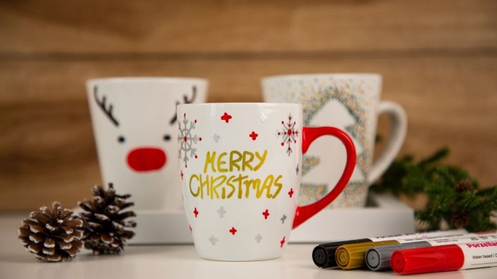 Eine bunte Tasse mit "MERRY CHRISTMAS" steht im Vordergrund, umgeben von weiteren Tassen und Bastelmaterialien.