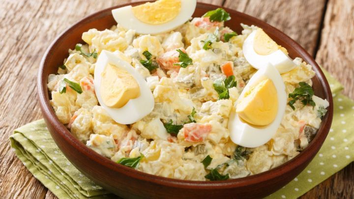 Ein goldbrauner Kartoffelsalat mit Speck und Mayo, garniert mit Eierscheiben, liegt in einer Holzsch&uuml;ssel auf einem rustikalen Tisch.