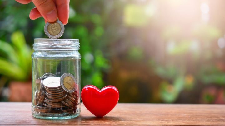 Eine Hand wirft eine M&uuml;nze in ein Glas mit bereits gesammelten M&uuml;nzen, daneben steht ein rotes Herzsymbol.