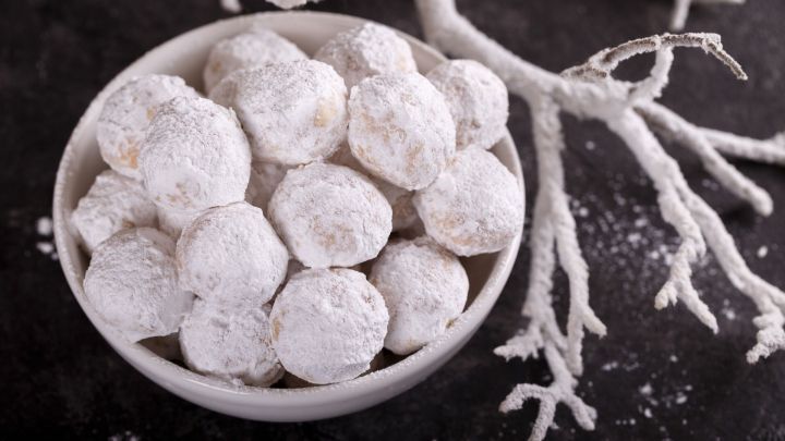 Weiche, runde Kourabiedes liegen in einer Sch&uuml;ssel, umgeben von zartem Puderzucker und einem frostigen Zweig.
