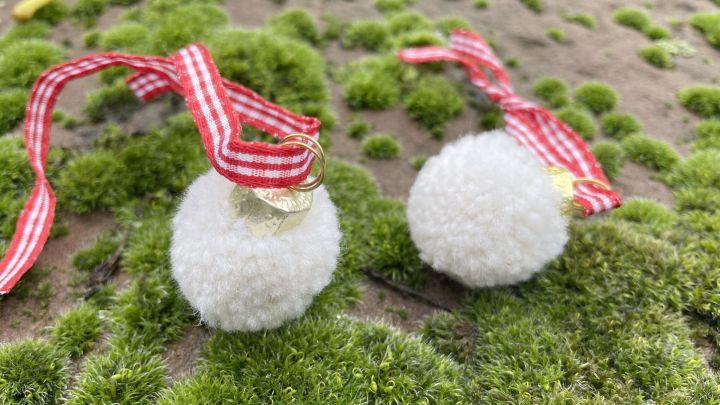 Zwei wei&szlig;e Pompom-Christbaumkugeln h&auml;ngen an roten Schleifen, platziert auf gr&uuml;nem Moosboden im Freien.