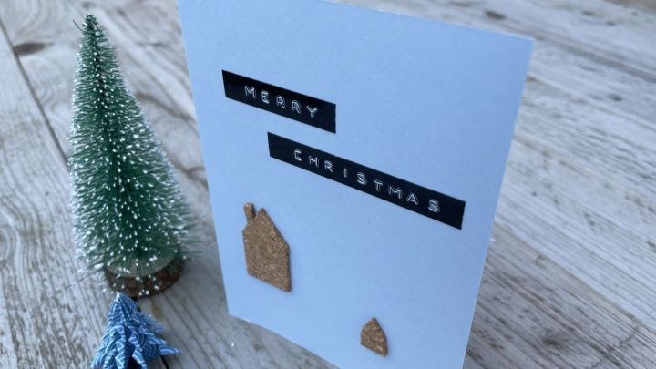 Eine Weihnachtskarte mit Korkh&auml;uschen steht auf einem Holztisch, dekoriert mit einem kleinen, gr&uuml;nen Baum. "MERRY CHRISTMAS" ist auf der Karte zu lesen.