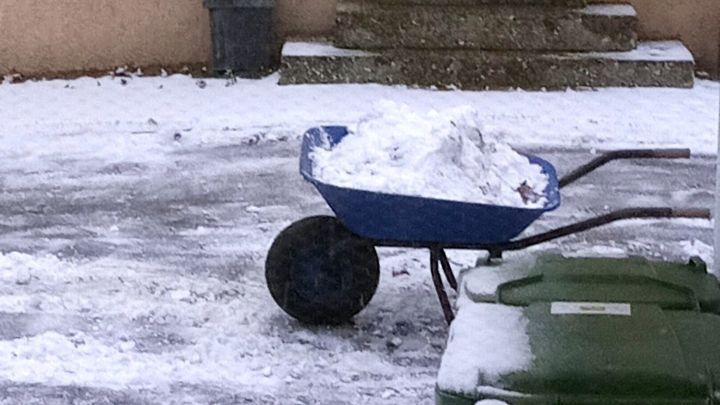 Eine Schubkarre voller Schnee wird auf einem schneebedeckten Au&szlig;enbereich neben M&uuml;lltonnen abtransportiert.