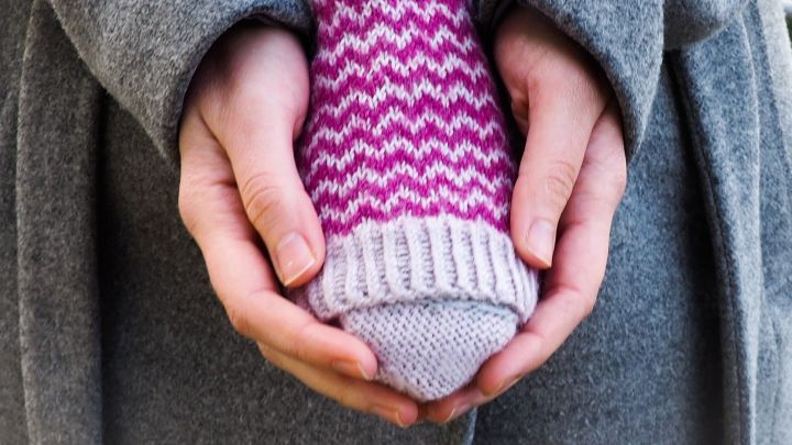 Ein gestrickter Handw&auml;rmer wird in den H&auml;nden einer Person gehalten, die einen grauen Mantel tr&auml;gt, um die K&auml;lte abzuwehren.