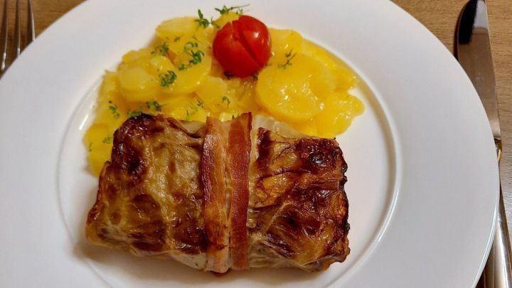 Ein saftiger Krautwickel mit Speck liegt auf einem wei&szlig;en Teller neben gelben Kartoffelscheiben und einer Kirschtomate.