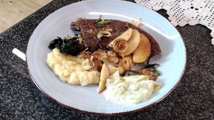 Ein gebratenes St&uuml;ck Leber liegt auf einem Teller, umgeben von Apfelst&uuml;cken, Zwiebeln, Brei und Gem&uuml;se.