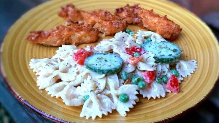 Ein bunter Nudelsalat aus Farfalle mit Erbsen, Gurken und Tomaten, serviert auf einem gelben Teller neben knusprigem H&auml;hnchen.