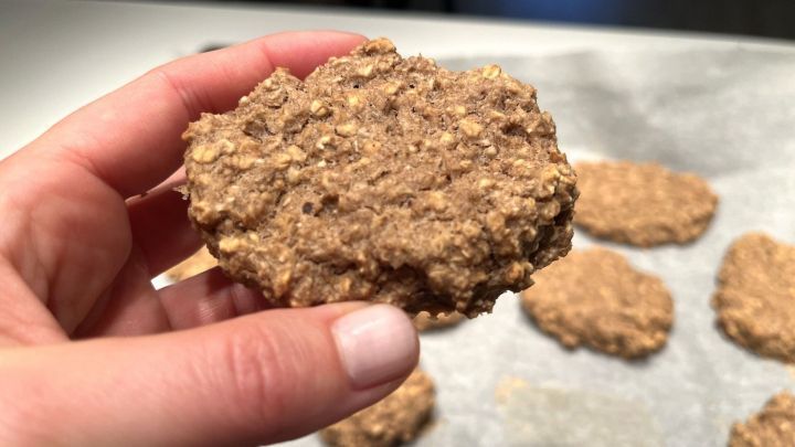Ein handgehaltener Fitness-Cookie aus Schokolade und Banane liegt vor einer Reihe &auml;hnlicher Kekse auf einem Backblech.