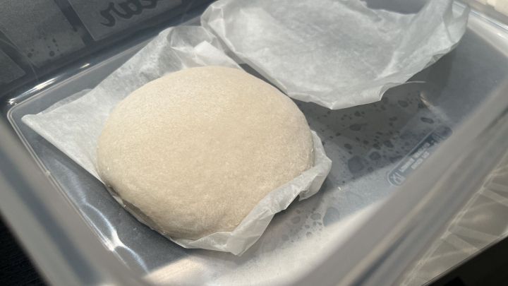 Ein runder Pizzateigling liegt auf Pergamentpapier in einem transparenten Beh&auml;lter, bereit zum Einfrieren.