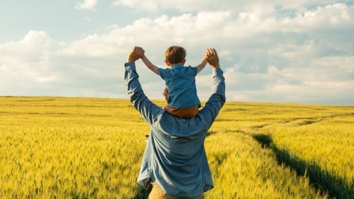 Ein Vater tr&auml;gt sein Kind auf den Schultern in einem goldenen Weizenfeld unter einem hellen Himmel.