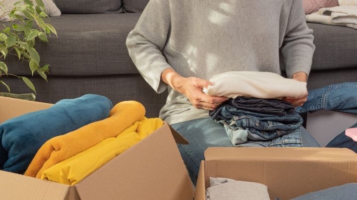 Eine Person faltet Kleidung und sortiert sie in Kartons, um minimalistisches Leben zu praktizieren. Ein heller Raum umgibt sie.