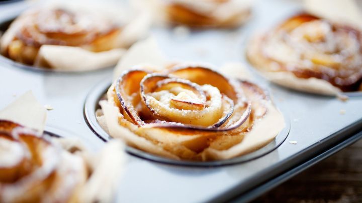 Bl&auml;tterteigrosen aus &Auml;pfeln liegen in einer Muffinform und sind leicht goldbraun, umgeben von Backpapier.