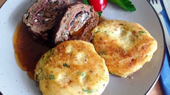 Saftige Hackfleisch-Roulade liegt auf einem Teller, umgeben von Kartoffel-Puffer und frischen Tomaten.