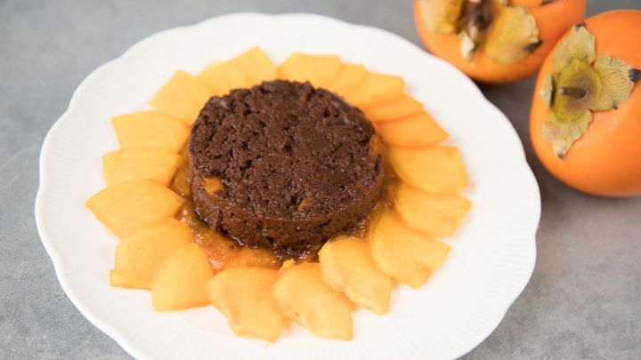 Schokoladiger Kaki-Pudding ist auf einem wei&szlig;en Teller angerichtet, umgeben von frischen Kaki-Scheiben.