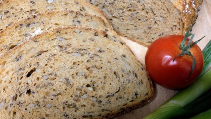 Ein frisch geschnittenes Fitnessbrot liegt auf einem Holzbrett, umgeben von einer Tomate und Fr&uuml;hlingszwiebeln.