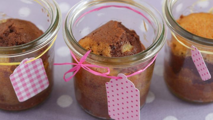 Marmorkuchen im Glas zeigt kleine Kuchen in Gl&auml;sern, verziert mit bunten B&auml;ndern und Etiketten, auf einem gepunkteten Hintergrund.