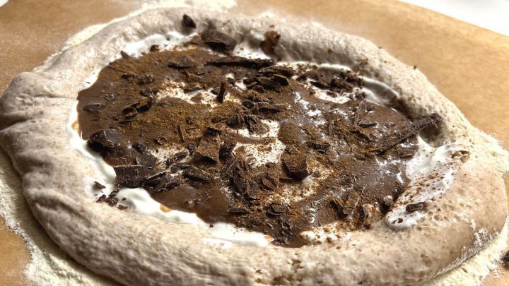 Eine Schokopizza mit Sauerteig liegt auf Backpapier und ist mit Schokolade und Creme gef&uuml;llt, umgeben von Mehl.