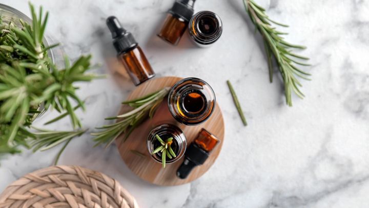 Braune Glasflaschen mit Rosmarin&ouml;l stehen auf einem Holzteller, umgeben von frischen Rosmarinzweigen auf einer wei&szlig;en Oberfl&auml;che.