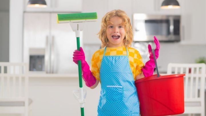 Ein Kind h&auml;lt einen Fensterreiniger und einen roten Eimer in einer modernen, hellen K&uuml;che und l&auml;chelt fr&ouml;hlich.