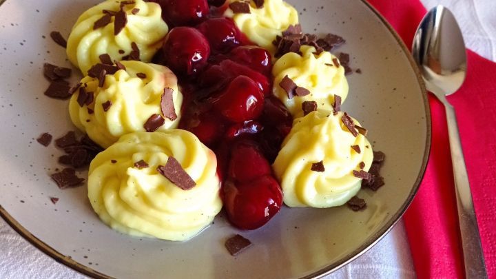 Zarte Vanillecremeh&auml;ufchen liegen in einer Sch&uuml;ssel, bedeckt mit hei&szlig;en Kirschen und Schokoladensp&auml;nen, auf hellem Tisch.