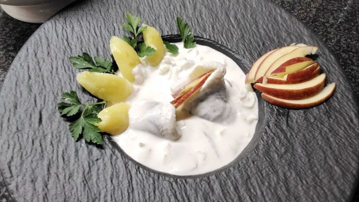 Marinierte Heringe liegen in einer wei&szlig;en Sauce, umgeben von Pellkartoffeln und Apfelscheiben auf einem dekorativen Teller.