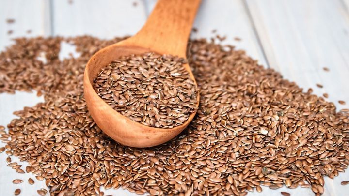 Braune Leinsamen liegen verstreut auf einem Holzuntergrund, w&auml;hrend ein Holzl&ouml;ffel sie aufnimmt, um als Magenschutz verwendet zu werden.