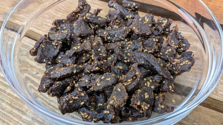 Dunkelbraunes Beef Jerky mit Sesam liegt in einer klaren Sch&uuml;ssel auf einem Holzuntergrund und wartet auf den Genuss.