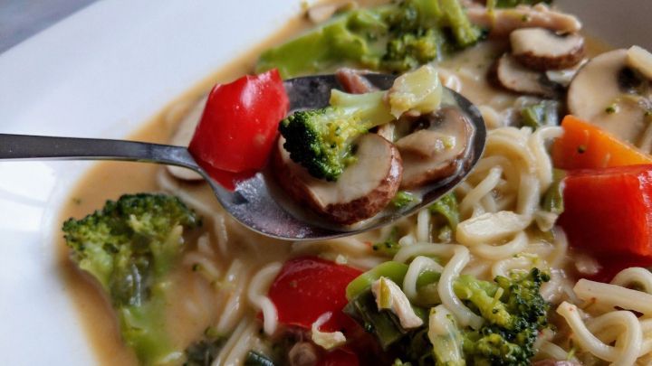 Eine L&ouml;ffel mit asiatischer H&uuml;hnersuppe hebt frisches Gem&uuml;se und Nudeln aus einer wei&szlig;en Sch&uuml;ssel voller Br&uuml;he.