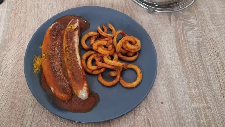 Auf einem blauen Teller liegen zwei Curryw&uuml;rste in So&szlig;e, neben knusprigen, spiralf&ouml;rmigen Pommes, auf einem Holztisch.