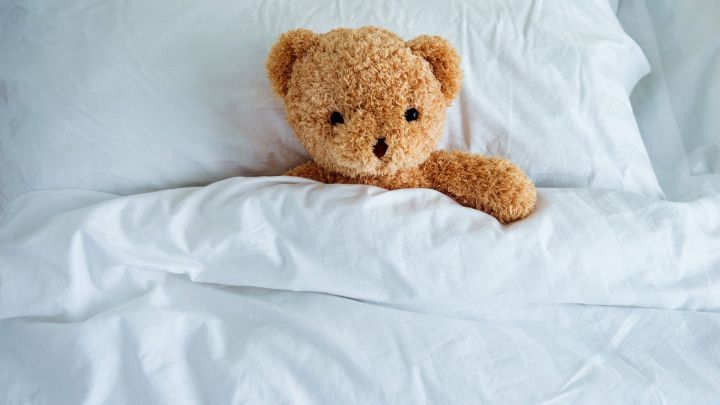Ein kuscheliger Teddyb&auml;r liegt unter der Bettdecke auf einem wei&szlig;en Kopfkissen, bereit zum Kuscheln.