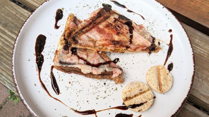 Zwei gegrillte K&auml;sesandwiches mit Bacon und einer k&ouml;stlichen Sauce sind auf einem wei&szlig;en Teller angerichtet, umgeben von Kr&auml;utern.