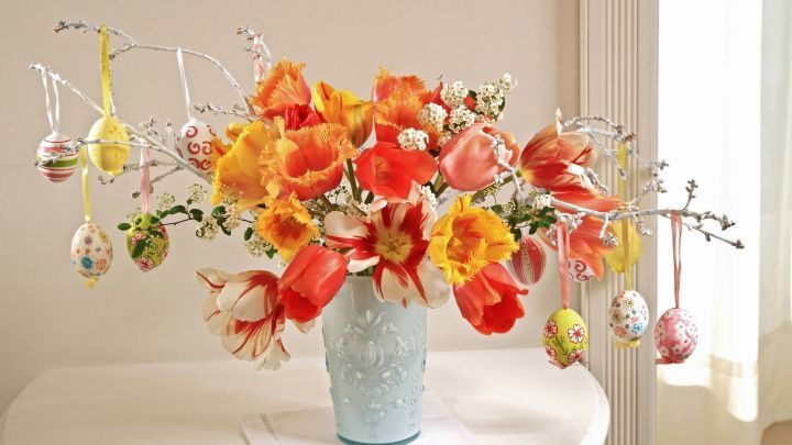 Bunte Osterzweige mit frischen Blumen in einer Vase stehen auf einem Tisch, umgeben von dekorativen Ostereiern.
