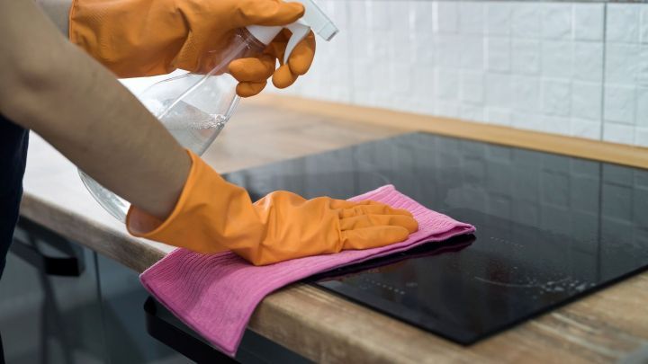 Eine Person reinigt ein Glaskeramikfeld mit einer Spr&uuml;hflasche und einem pinken Tuch in einer modernen K&uuml;che.