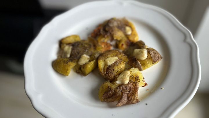 Goldbraun gebratene Quetschkartoffeln liegen auf einem wei&szlig;en Teller, garniert mit einer hellen Sauce und serviert im Innenraum.