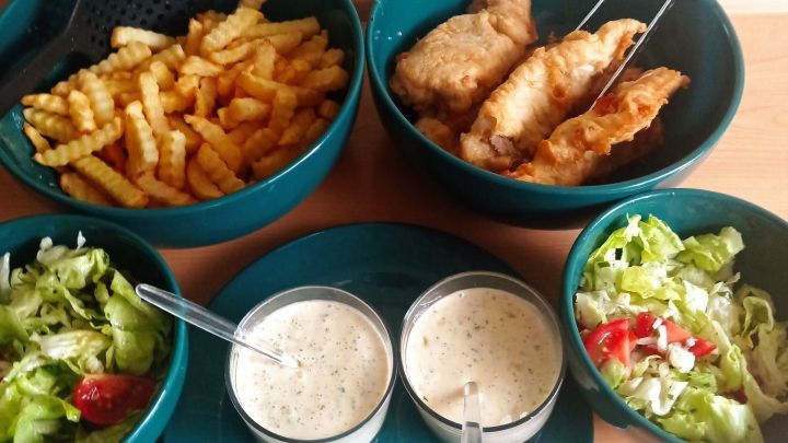 Frittierter Fisch liegt neben knusprigen Pommes und frischem Salat in bunten Schalen auf einem Holzbrett.