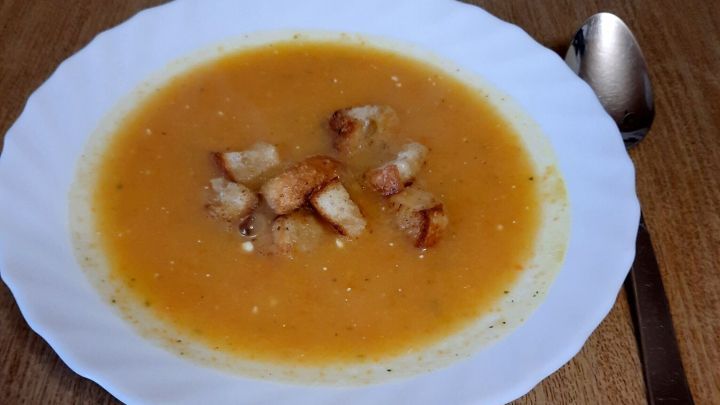 Eine cremige Wurzelgem&uuml;se-Suppe in einem wei&szlig;en Teller, garniert mit knusprigen Brotw&uuml;rfeln, steht auf einem Holztisch.