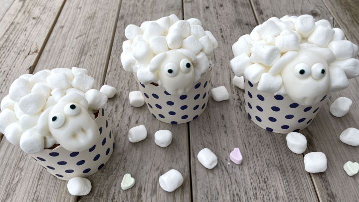 Drei Muffins, dekoriert mit Marshmallows, sehen aus wie Sch&auml;fchen und stehen auf einer Holzoberfl&auml;che.