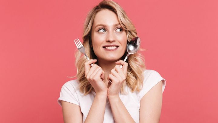 Eine l&auml;chelnde Frau h&auml;lt eine Gabel und einen L&ouml;ffel, umgeben von einem rosa Hintergrund, was auf gesunde Ern&auml;hrung hinweist.