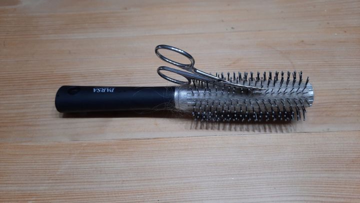 Eine Rundb&uuml;rste mit Haaren und einer Nagelschere liegt auf einem hellen Holztisch.