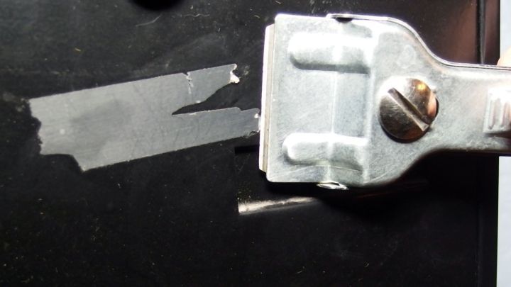 Ein Briefkasten mit Kleberesten wird mit einem Schaber gereinigt. Die Umgebung ist dunkel und zeigt metallische Details.