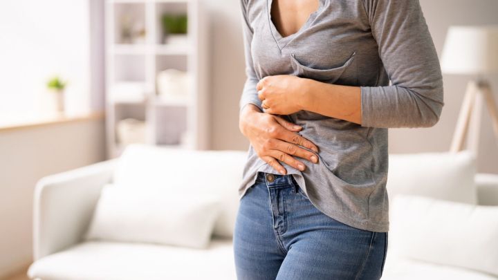 Eine Person mit Bauchschmerzen h&auml;lt sich den Bauch in einem hellen Raum mit einem Sofa, was auf Sodbrennen oder Gastritis hindeutet.