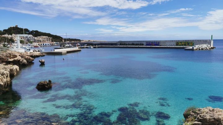 Kristallklares Wasser umsp&uuml;lt die felsige K&uuml;ste, w&auml;hrend Boote im Hafen anlegen, ideal f&uuml;r Urlaubstipps und Ersparnisse.