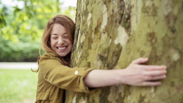 Eine Frau umarmt einen Baum im Freien mit einem gl&uuml;cklichen L&auml;cheln, was ein Zeichen f&uuml;r umweltfreundliches Leben darstellt.