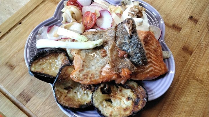 Auf dem Teller liegt gegrillter Lachs mit gebratenem Gem&uuml;se und frischem Salat, angerichtet auf einem Holzuntergrund.