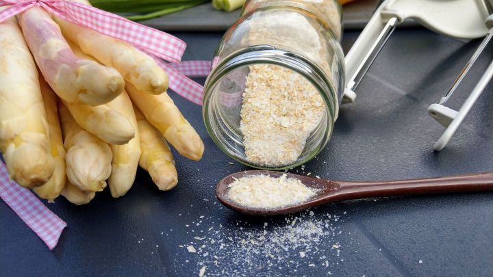 Frischer Spargel liegt mit einem Glas Spargelsalz daneben auf einem Tisch, w&auml;hrend etwas Salz auf einen L&ouml;ffel gefallen ist.