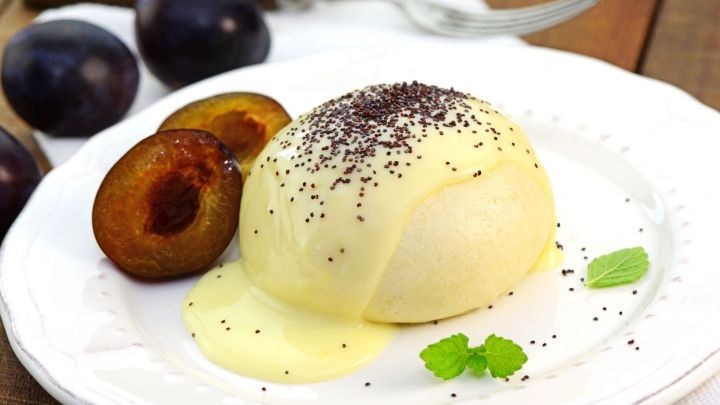 Eine Dampfnudel ohne Hefe liegt auf einem Teller, umgeben von Pflaumen und einer s&uuml;&szlig;en So&szlig;e, in einem rustikalen Ambiente.