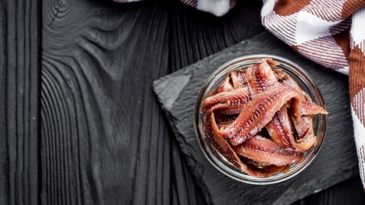 Ein Glas mit Sardellen steht auf einem schwarzen Brett, umgeben von einer karierten Serviette und einem Holzuntergrund.