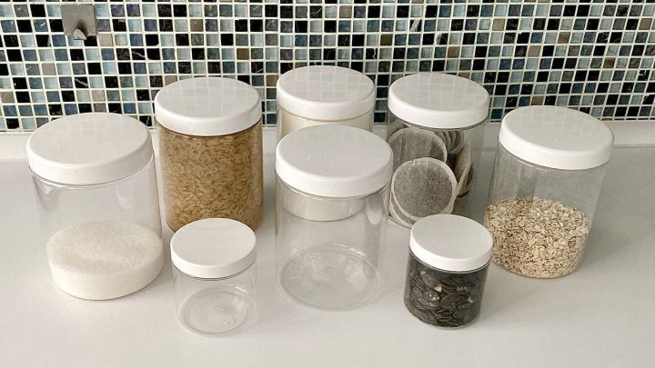 Beholder mit verschiedenen Lebensmitteln wie Reis, Haferflocken und Zucker werden ordentlich auf einer wei&szlig;en Oberfl&auml;che vor einer bunten Fliesenwand angeordnet.