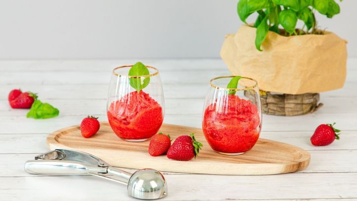 Zwei Gl&auml;ser mit frischem Erdbeersorbet stehen auf einem Holzbrett, umgeben von Erdbeeren und einem Basilikumtopf.